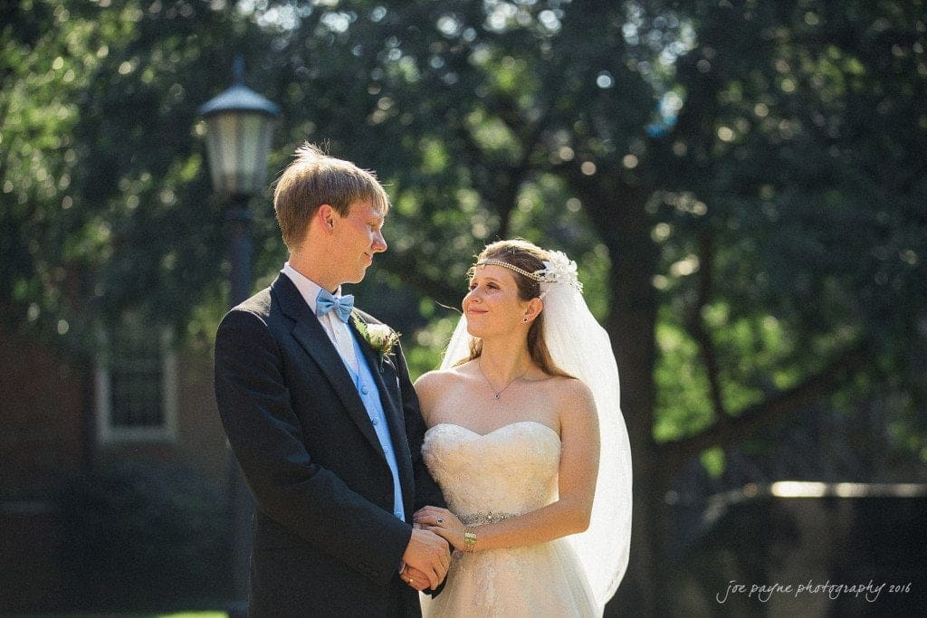 duke chapel & carolina inn wedding - sidney & andrew A bride and groom stand outdoors, gazing at each other lovingly. The bride wears a strapless white gown and veil, and the groom is in a black suit with a blue bow tie. They are holding hands, with trees and a lamppost in the background.