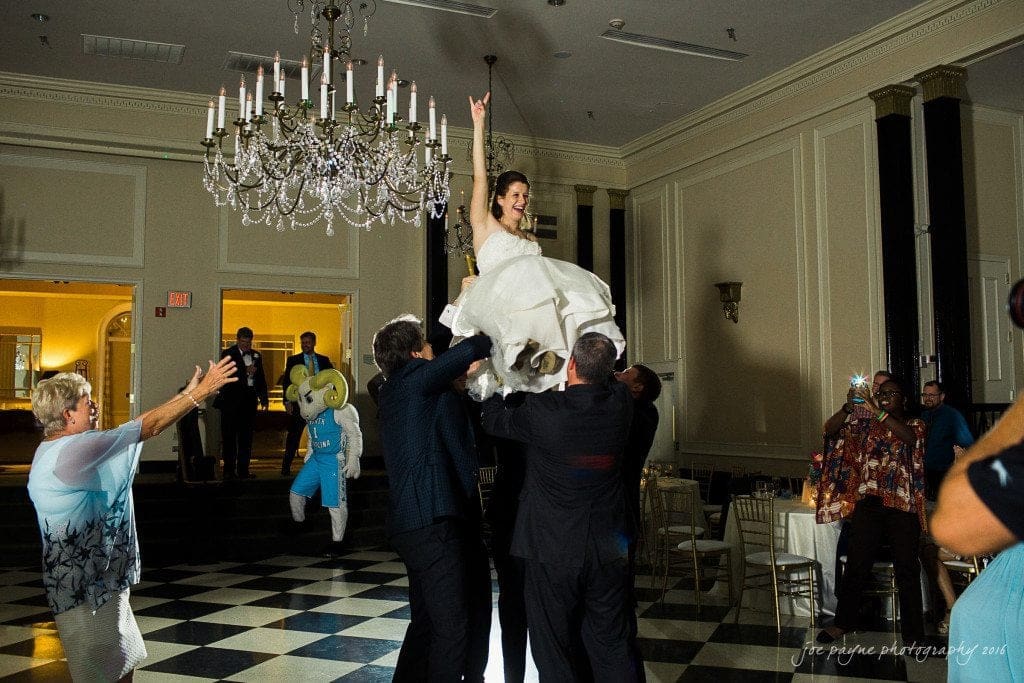 duke chapel & carolina inn wedding - sidney & andrew A bride is lifted into the air by several people in a ballroom with a black and white checkered floor and a chandelier. Guests cheer and a person in a blue and white mascot costume stands in the background.