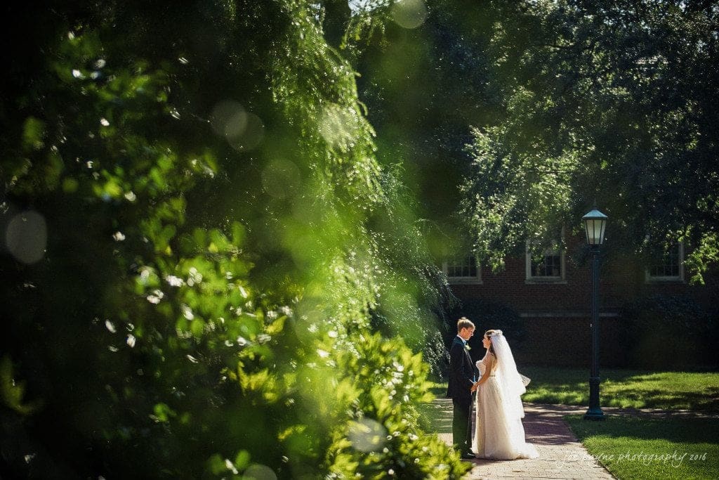 duke chapel & carolina inn wedding - sidney & andrew A bride and groom stand together on a sunny pathway, surrounded by lush green foliage and illuminated by soft, dappled sunlight. The background features trees and a brick building.