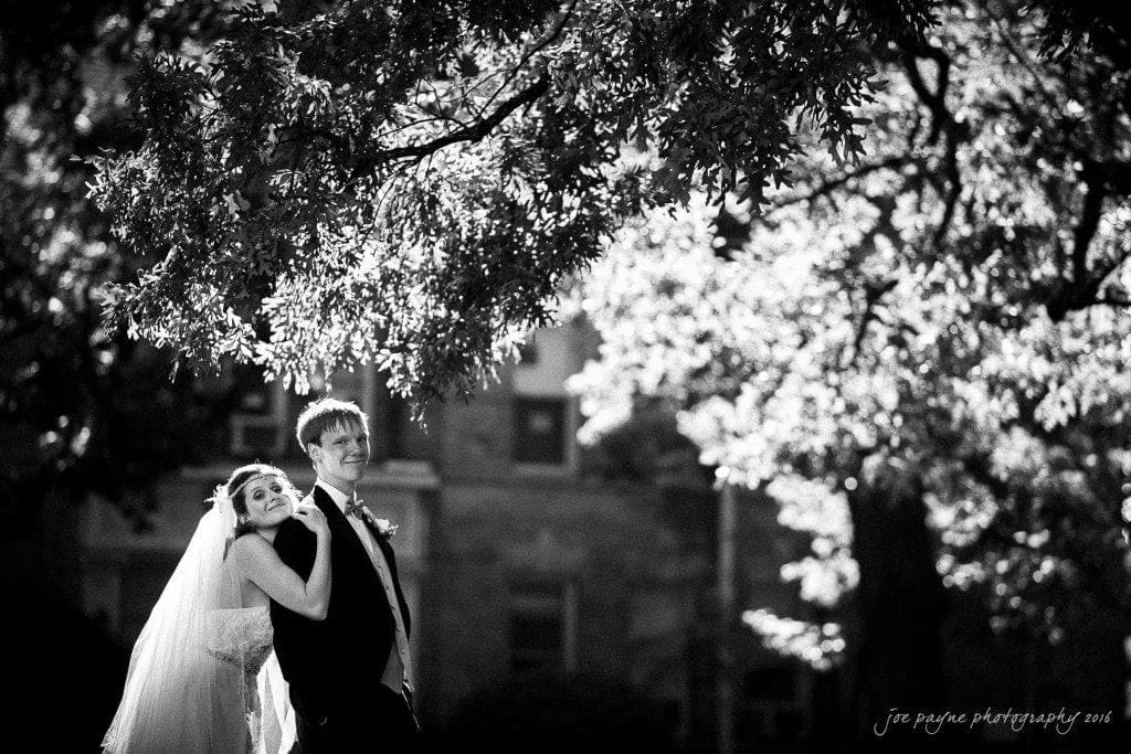 duke chapel & carolina inn wedding - sidney & andrew A black and white photo of a bride and groom smiling under a large tree. The bride rests her head on the grooms shoulder, with a veil trailing behind her. Sunlight filters through the leaves. A building is visible in the background.