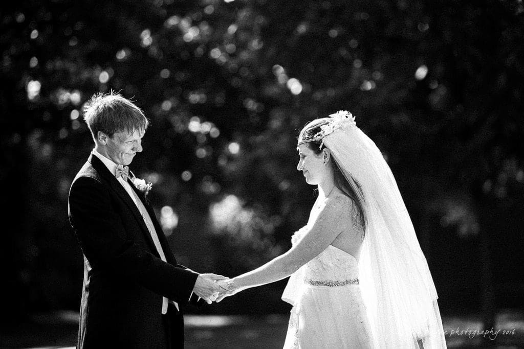 duke chapel & carolina inn wedding - sidney & andrew A black-and-white photo of a couple holding hands at an outdoor wedding. The bride wears a long veil and dress, and the groom is in a suit with a bow tie. They are looking at each other against a backdrop of trees.