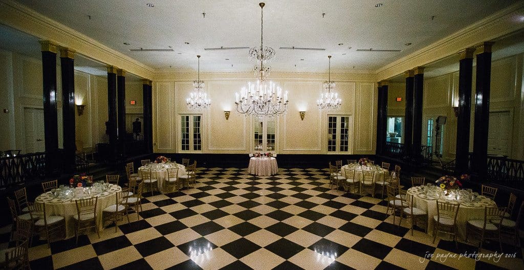 duke chapel & carolina inn wedding - sidney & andrew A spacious ballroom with a checkered floor is elegantly set for an event. Six round tables with floral centerpieces and gold chairs surround a central table. Two large chandeliers hang from the ceiling, illuminating the room.