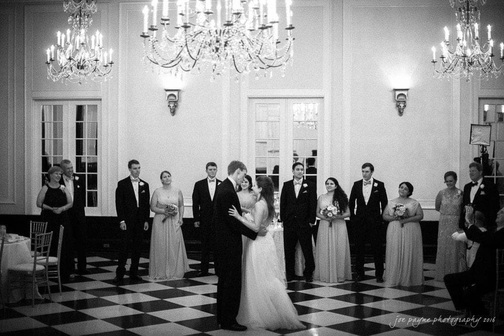 duke chapel & carolina inn wedding - sidney & andrew A bride and groom share their first dance in a grand ballroom with chandeliers. Bridesmaids and groomsmen stand nearby watching. The floor is checkered, and the setting exudes a classic, elegant ambiance.