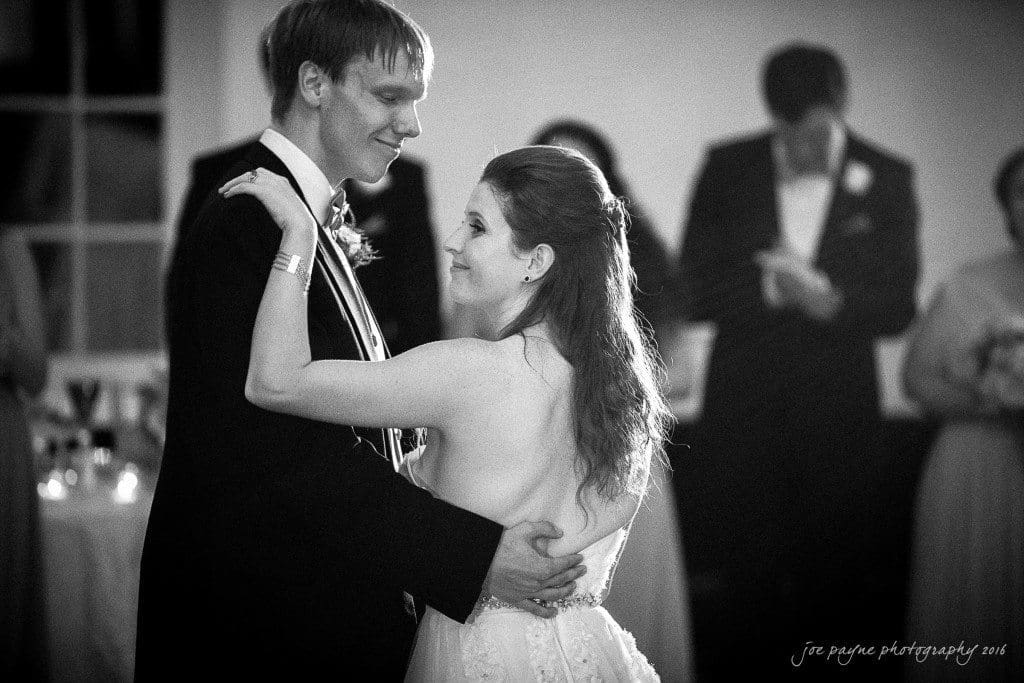 duke chapel & carolina inn wedding - sidney & andrew A couple shares their first dance at a wedding reception. They smile at each other, surrounded by blurred guests in the background. The scene is warmly lit, creating an intimate and joyful atmosphere.