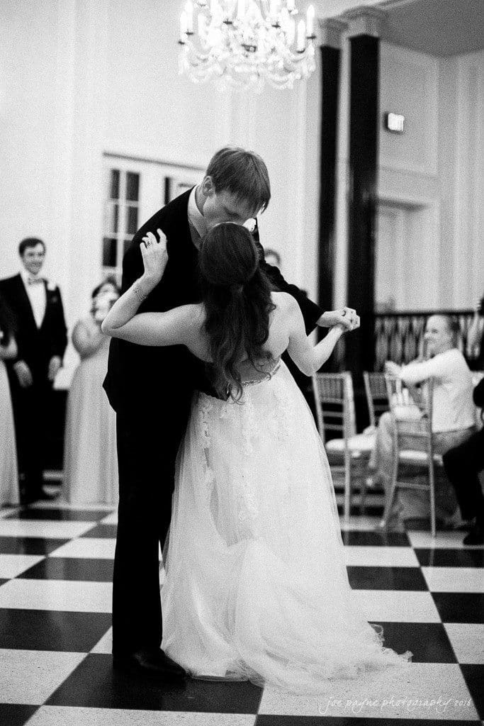 duke chapel & carolina inn wedding - sidney & andrew A black and white photo of a couple dancing on a checkered floor. The man, in a suit, leans in to kiss the woman, who wears a flowing wedding dress. They are surrounded by elegantly dressed guests seated and standing, watching the moment.