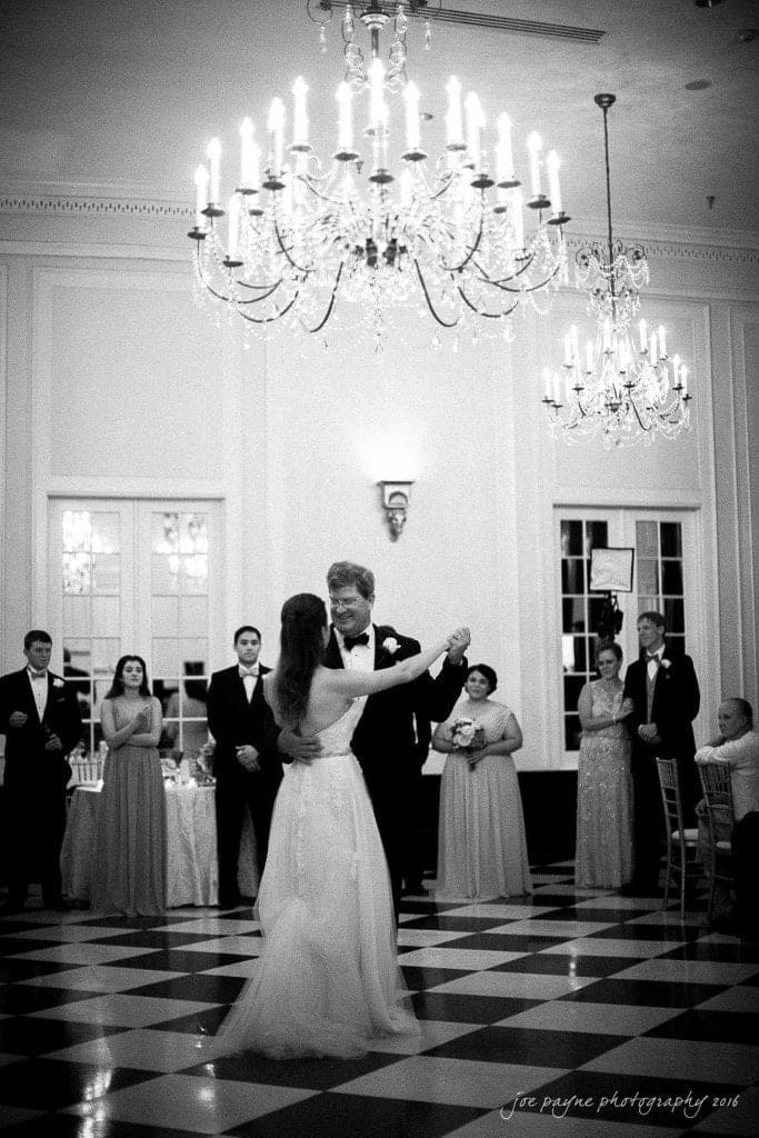 duke chapel & carolina inn wedding - sidney & andrew A bride and groom share their first dance in a grand ballroom, surrounded by chandeliers and guests in formal attire. The black and white image captures an elegant and romantic atmosphere.