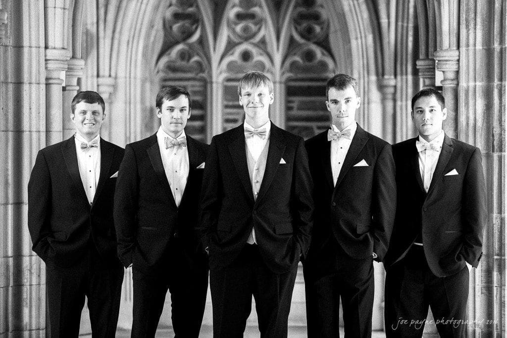 duke chapel & carolina inn wedding - sidney & andrew Five men in tuxedos and bow ties stand in front of an ornate stone archway. They are facing the camera with neutral expressions. The setting appears formal, possibly a wedding or event. Black and white image.