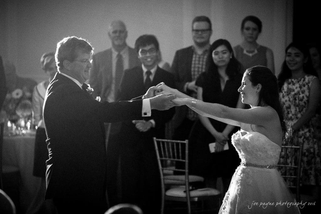duke chapel & carolina inn wedding - sidney & andrew A bride and an older man, perhaps her father, are dancing in formal attire at a wedding reception. They are surrounded by smiling onlookers, including men and women in suits and dresses. The setting is elegant, with dim lighting.