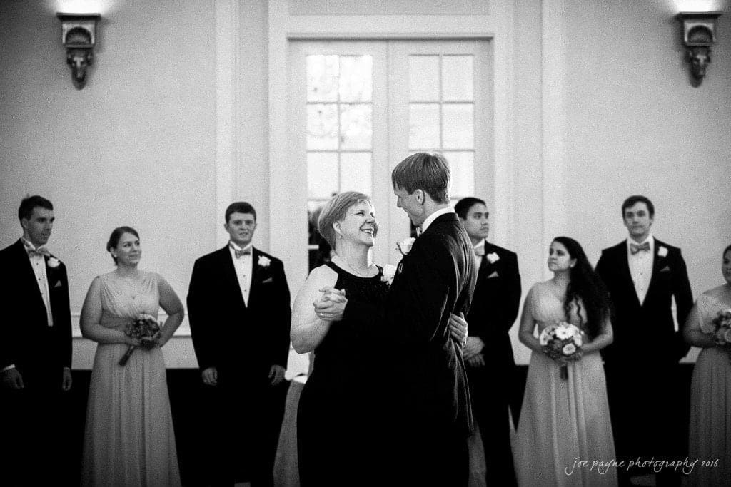 duke chapel & carolina inn wedding - sidney & andrew A black and white photo of a couple dancing together inside a room with a group of people in formal attire standing around them, watching. The woman is wearing a dark dress, and the man is in a suit with a bow tie.