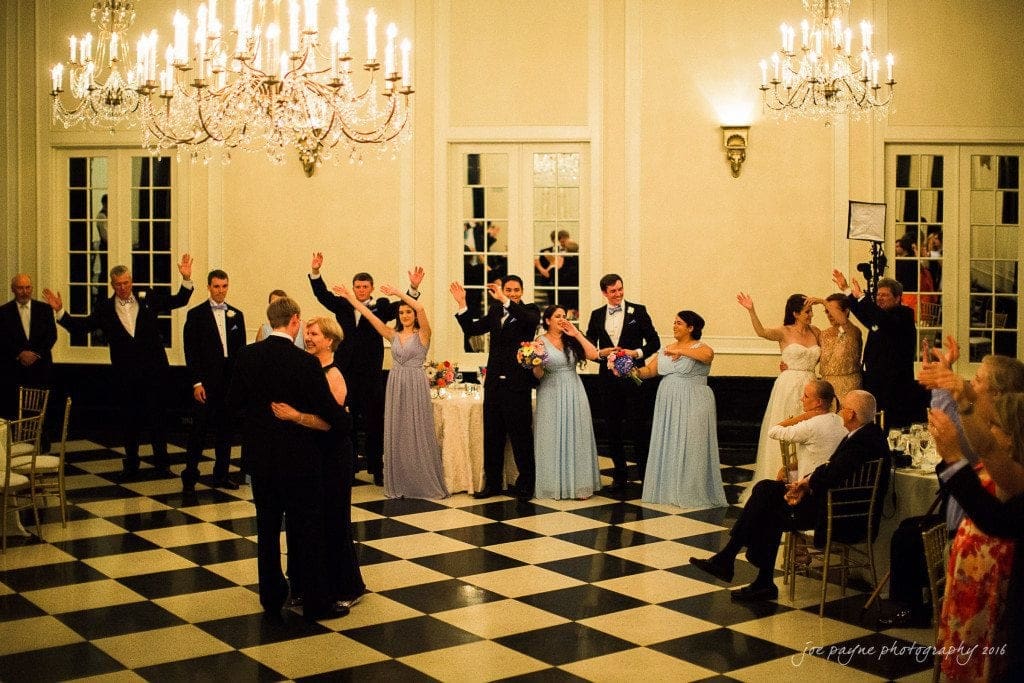 duke chapel & carolina inn wedding - sidney & andrew A couple dances under chandeliers on a black-and-white checkered floor. Guests, some dressed in formal attire and holding flowers, stand in a line, clapping and cheering, in a room with large windows and elegant lighting.