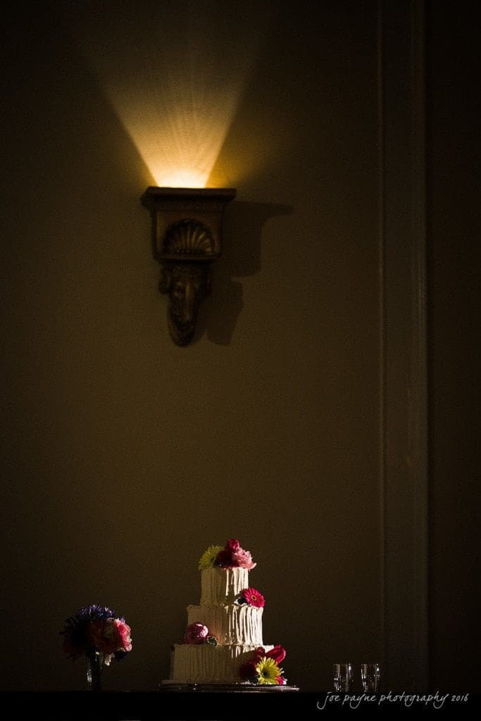 duke chapel & carolina inn wedding - sidney & andrew A tiered white wedding cake decorated with pink and red flowers is on a table under a sconce light. Nearby are two glasses and a bouquet of colorful flowers. The background is dimly lit, highlighting the cake.