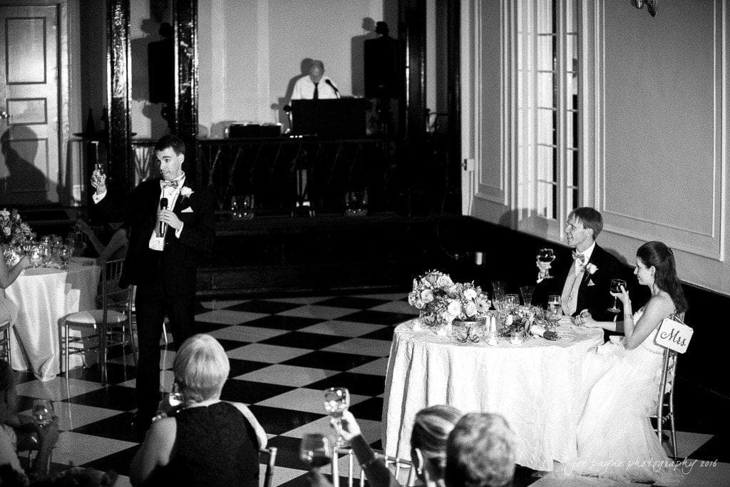 duke chapel & carolina inn wedding - sidney & andrew A black and white photo of a formal celebration. A man in a suit stands raising a toast, while a seated couple, labeled Mr. and Mrs., smile at him. Guests seated around tables and a person at a DJ booth are visible in the background.