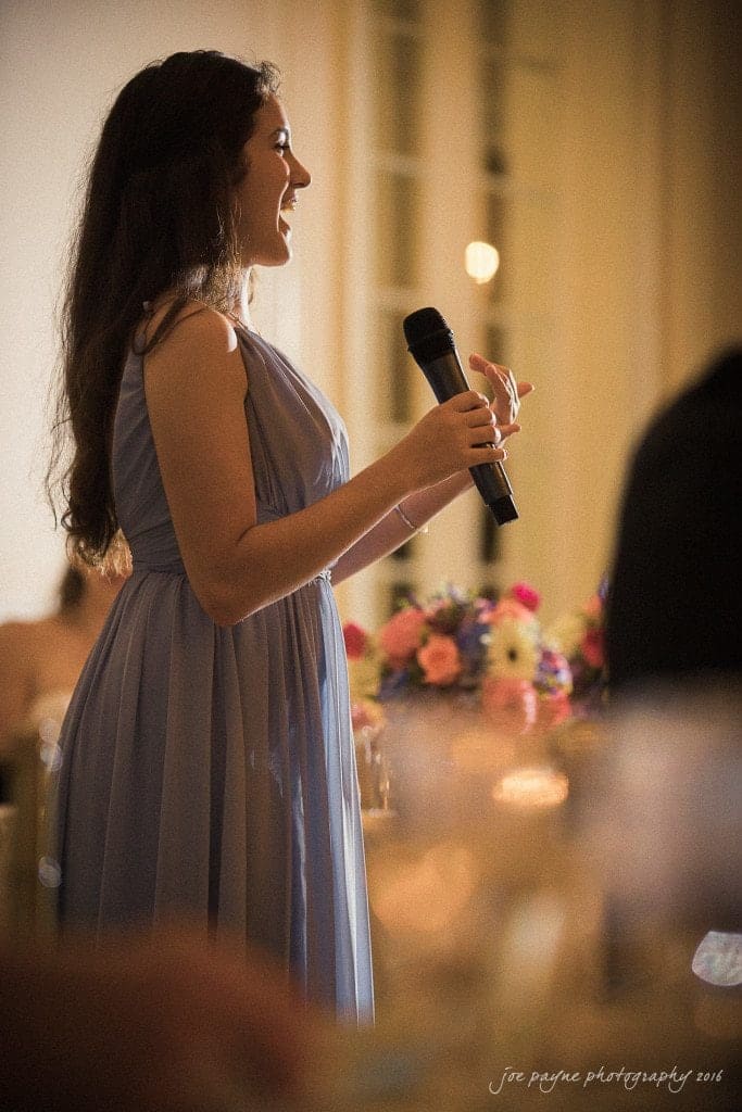 duke chapel & carolina inn wedding - sidney & andrew A woman in a lavender dress holds a microphone while speaking at an indoor event. She stands near a table with a colorful floral centerpiece, and soft lighting creates a warm atmosphere in the room.