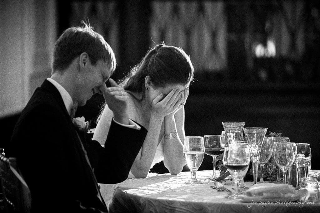 duke chapel & carolina inn wedding - sidney & andrew A bride and groom sit at a table, sharing a moment of laughter. The groom covers his face with his hand while the bride holds her hands to her face, smiling. Various glasses and tableware are in front of them. The scene is in black and white.