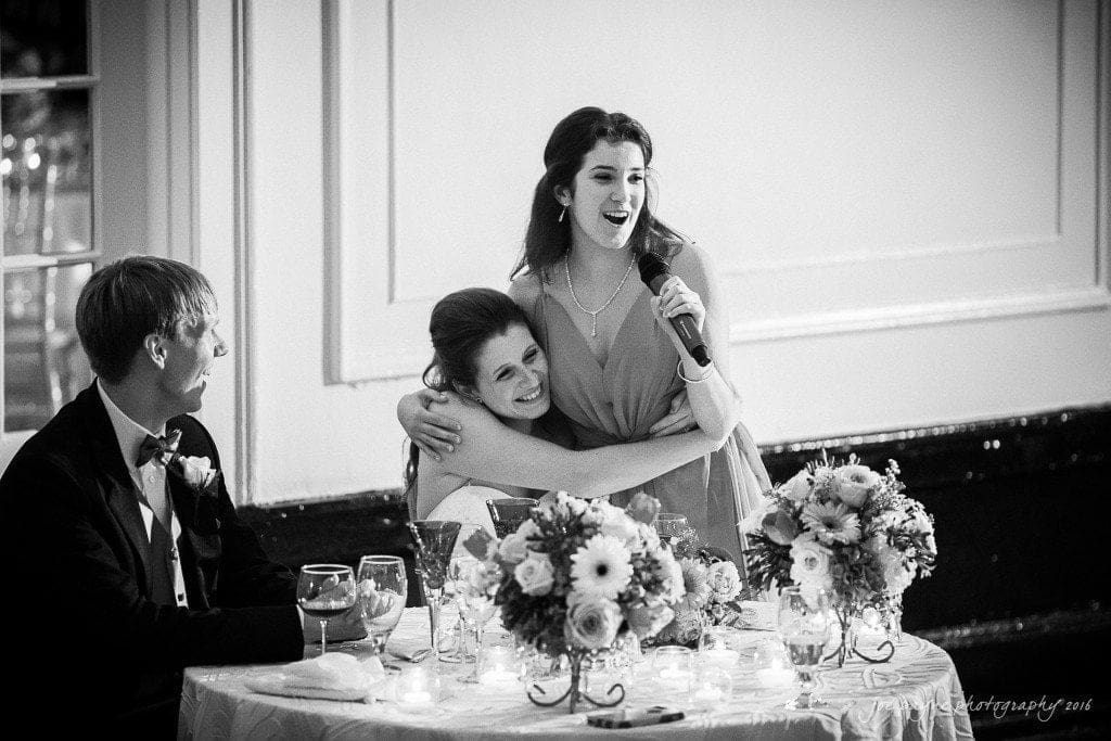 duke chapel & carolina inn wedding - sidney & andrew A joyful wedding scene with a bridesmaid hugging the bride while speaking into a microphone. The groom smiles beside them. The table is elegantly set, adorned with flowers and wine glasses.