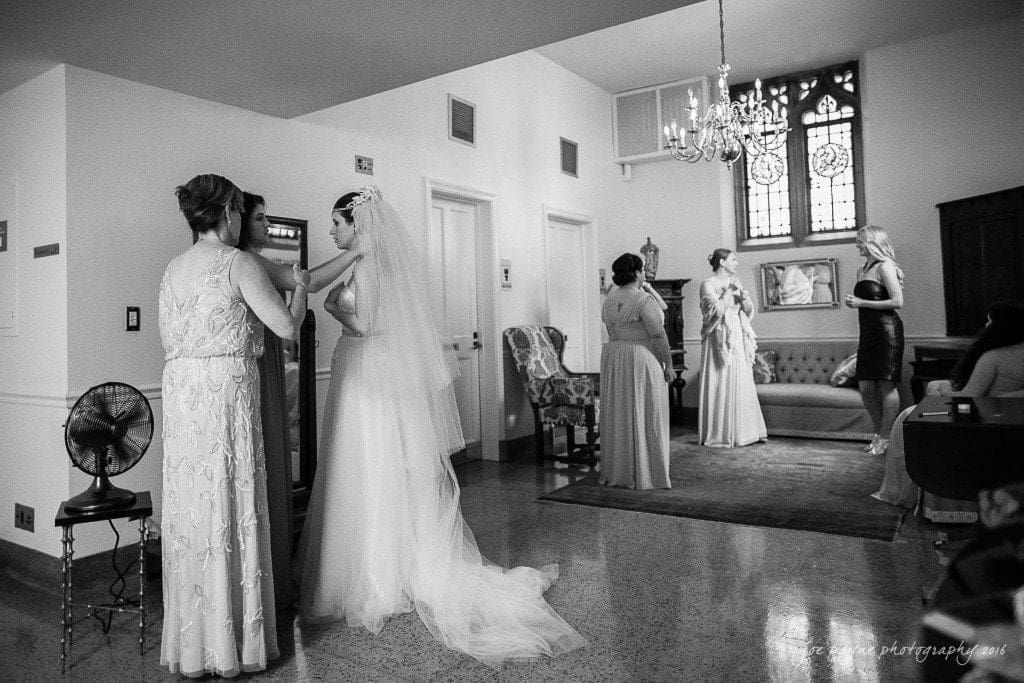 duke chapel & carolina inn wedding - sidney & andrew A black and white photo of a bridal party getting ready in a room. The bride, in a long dress with a veil, is assisted by a woman. Others sip drinks and adjust dresses. A chandelier and ornate window add elegance to the setting.
