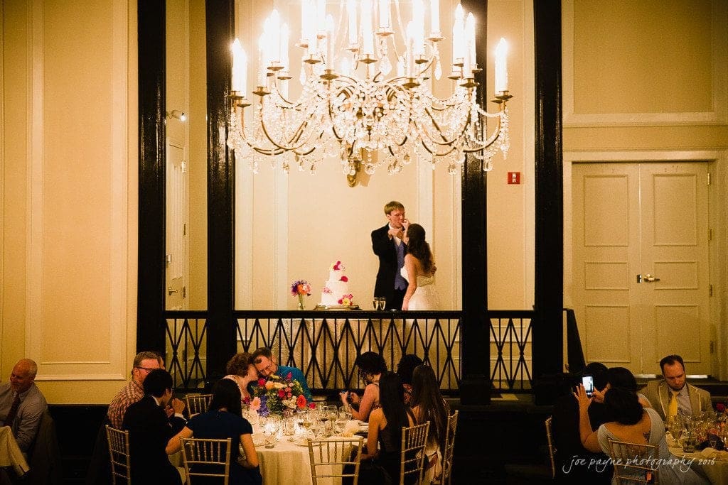 duke chapel & carolina inn wedding - sidney & andrew A couple stands in front of a large cake, sharing a kiss under an elegant chandelier at a wedding reception. Guests are seated at tables, capturing the moment with their phones. The room is warmly lit and decorated with flowers.