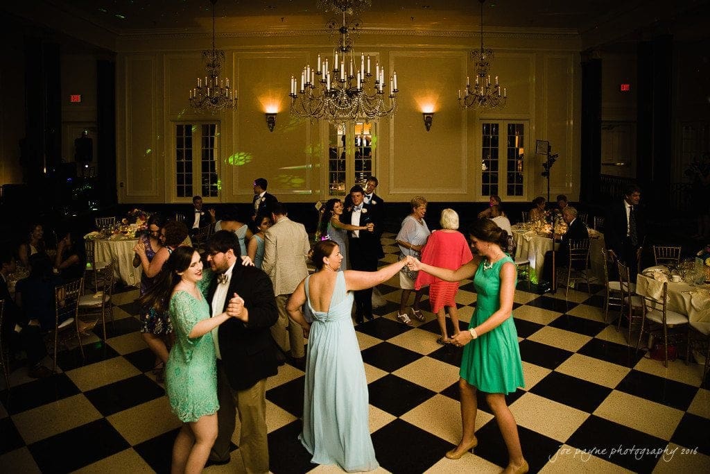 duke chapel & carolina inn wedding - sidney & andrew Guests dance on a black and white checkered floor under chandeliers in a formal event hall. People are dressed in semi-formal attire, and tables are elegantly set in the background.