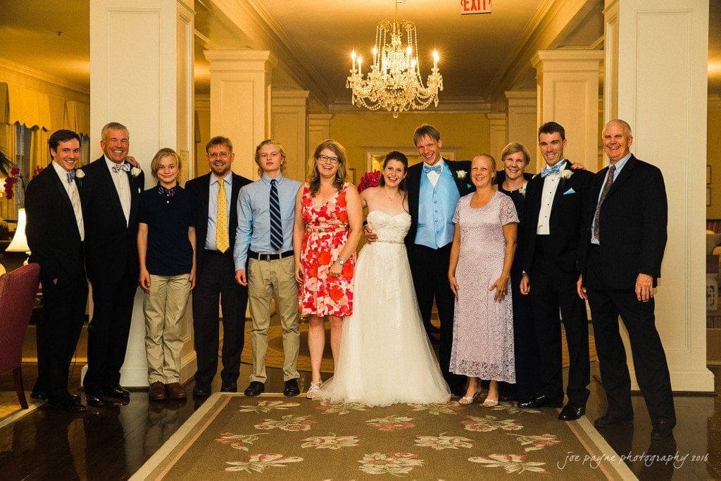 duke chapel & carolina inn wedding - sidney & andrew A group of people stands in a well-lit room beneath a chandelier. The bride in a white gown and a groom in a light blue vest are at the center. Others are dressed in formal and semi-formal attire, posing together and smiling.