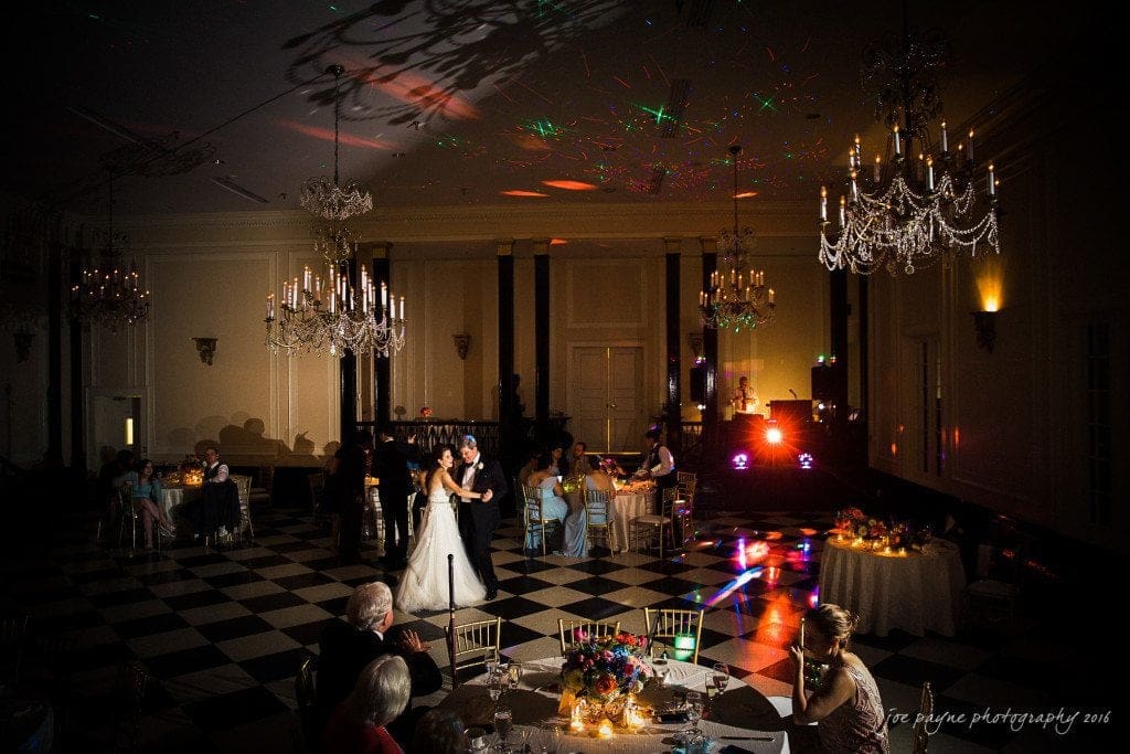 duke chapel & carolina inn wedding - sidney & andrew A wedding reception in an elegant ballroom with chandeliers, where a bride and groom are dancing on a checkered floor. Guests are seated at round tables decorated with flowers and lit candles. Colorful lights illuminate the room.
