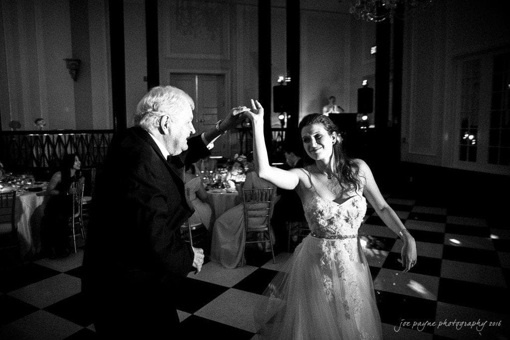 duke chapel & carolina inn wedding - sidney & andrew A bride in a flowing dress dances joyfully with an older gentleman in a suit on a checkered dance floor at a formal event. The room is elegantly decorated, with guests seated at tables in the background.