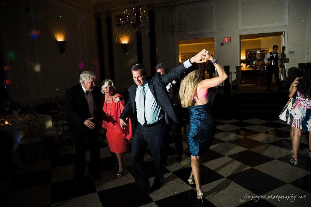 duke chapel & carolina inn wedding - sidney & andrew A group of people dancing in a dimly lit room with checkered flooring. A man in a suit twirls a woman in a blue dress. Other guests, dressed in formal attire, watch and smile. Chandeliers and a lit exit sign are visible in the background.