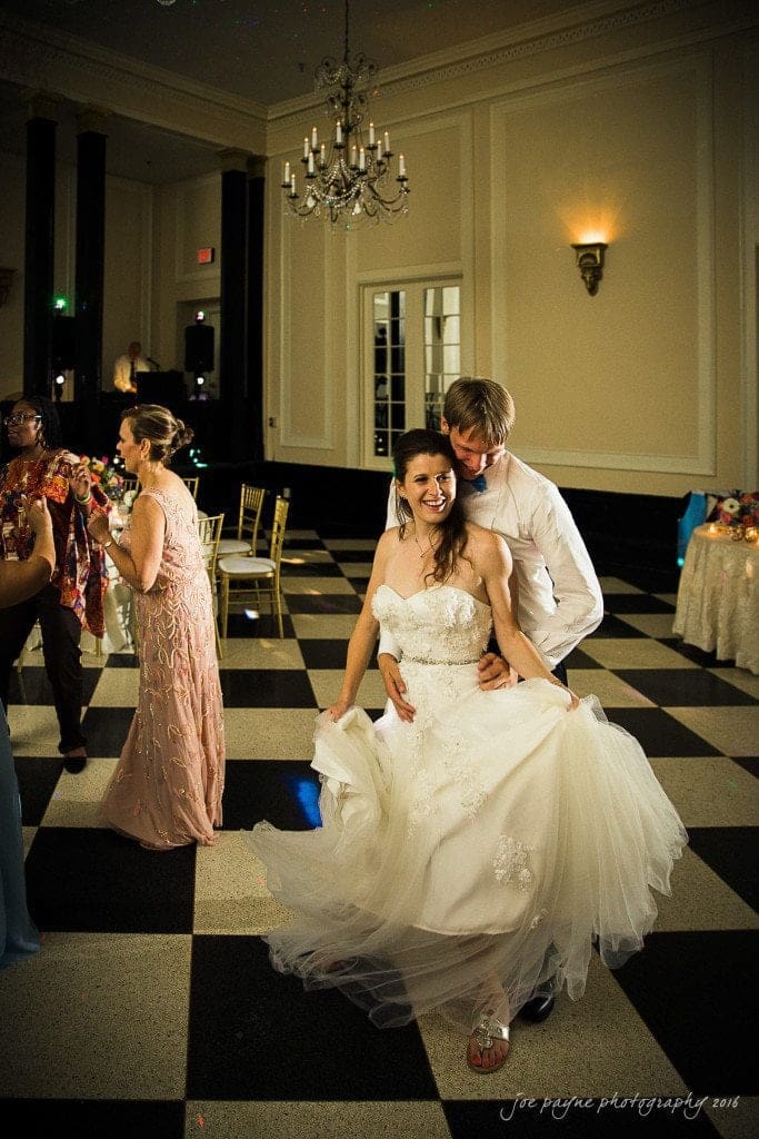 duke chapel & carolina inn wedding - sidney & andrew A bride and groom dance joyfully in a ballroom with black and white checkered flooring. Other guests are seen dancing in the background. The room is elegantly decorated with chandeliers and wall sconces.