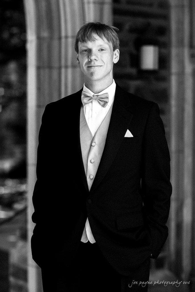 duke chapel & carolina inn wedding - sidney & andrew A man in a tuxedo stands confidently in a black and white photo. He wears a bow tie and vest, with hands in pockets. The background features arched architecture.