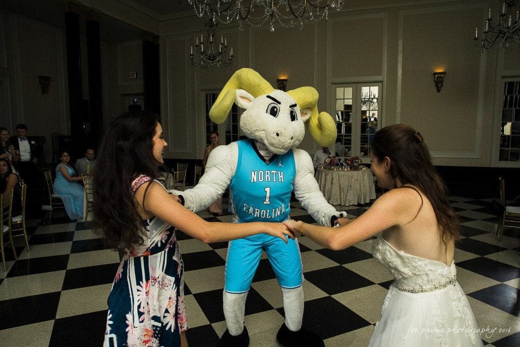 duke chapel & carolina inn wedding - sidney & andrew A group of people are dancing with a person in a North Carolina ram mascot costume at an indoor event. The room features checkered flooring, chandeliers, and finely dressed guests sitting at round tables.