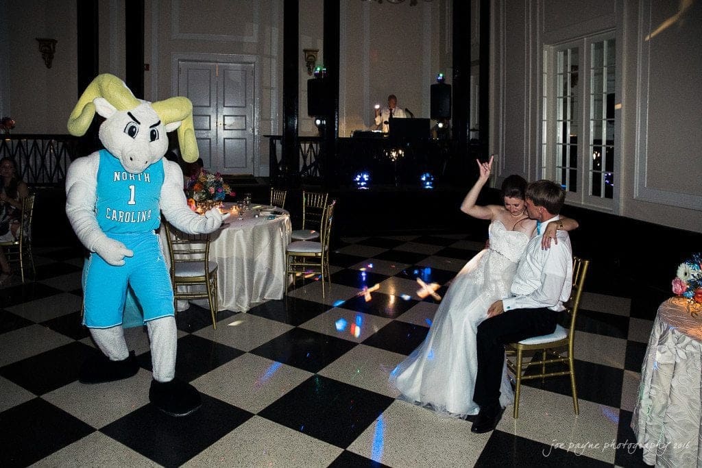 duke chapel & carolina inn wedding - sidney & andrew A wedding reception with a couple seated on a chair. A person in a North Carolina mascot costume is dancing nearby. The woman raises her hand, smiling. The checkered floor and a dressed-up DJ booth are visible in the background.