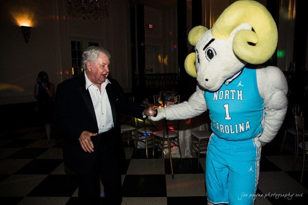 duke chapel & carolina inn wedding - sidney & andrew An elderly man in formal attire playfully interacts with a person in a North Carolina Tar Heels mascot costume. They are in a dimly lit room with checkered flooring and decorative seating.