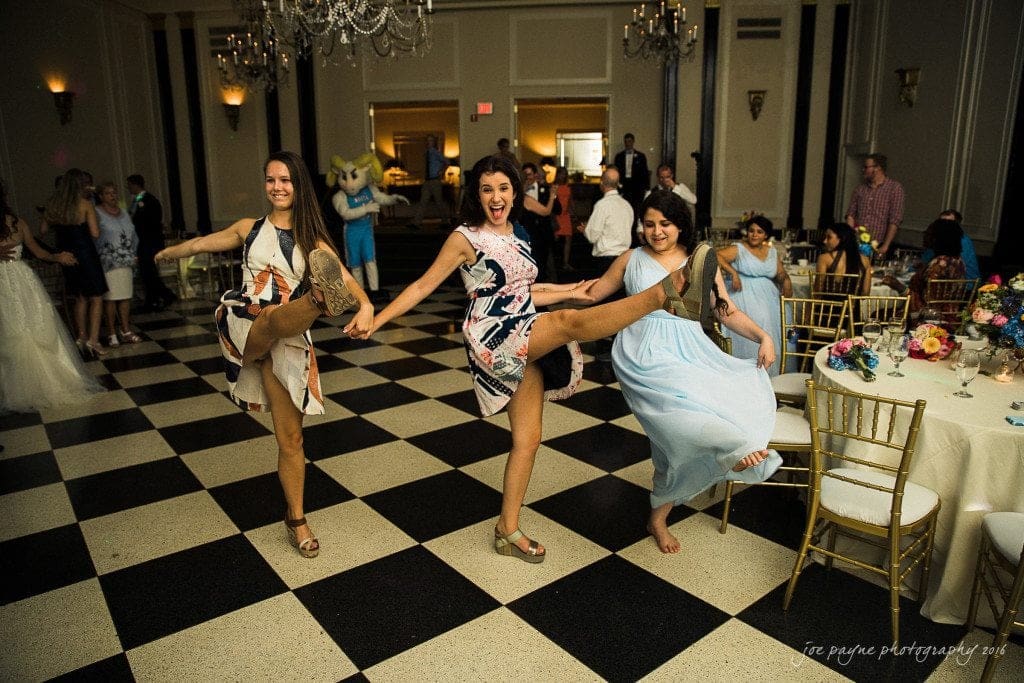 duke chapel & carolina inn wedding - sidney & andrew Three women in colorful dresses joyfully kick their legs up while dancing on a black and white checkered floor at a lively event. Guests and tables with floral centerpieces surround them, adding to the festive atmosphere.