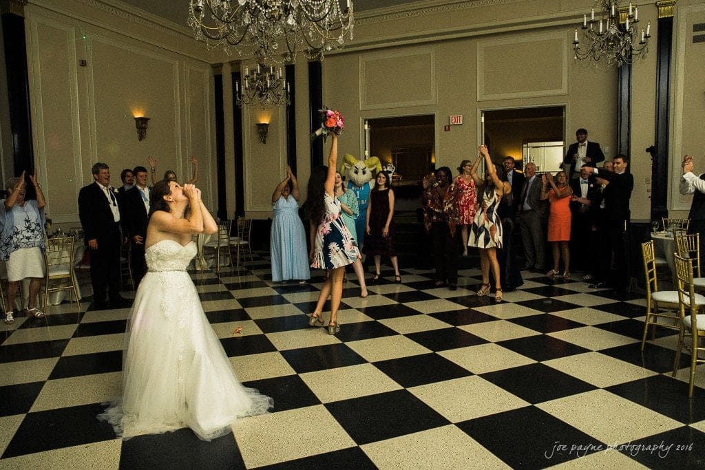 duke chapel & carolina inn wedding - sidney & andrew A bride in a white wedding dress is tossing her bouquet in a ballroom. Guests cheer and reach up to catch it. The room has black and white checkered flooring and elegant chandeliers.