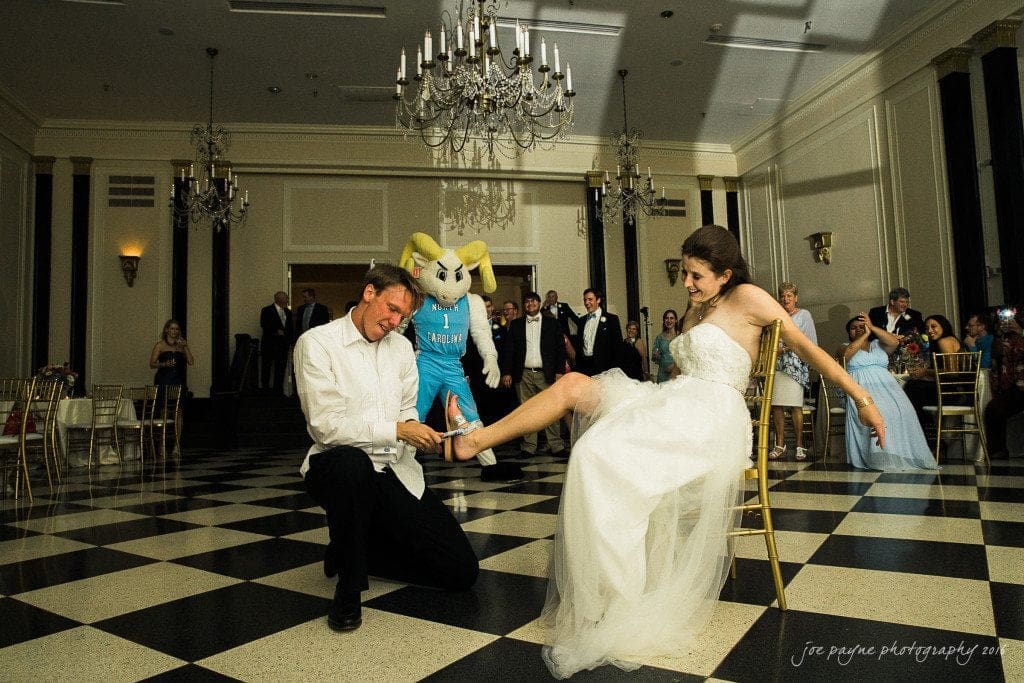 duke chapel & carolina inn wedding - sidney & andrew A bride sits on a chair as the groom kneels and removes her garter in a ballroom. The couple is surrounded by guests, and a person in a bunny costume stands behind them. Chandeliers hang from the ceiling.