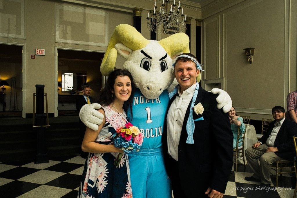 duke chapel & carolina inn wedding - sidney & andrew A smiling couple stands with a large ram mascot wearing a North Carolina jersey. The woman holds a colorful bouquet, and the man wears a suit with a light blue tie wrapped humorously around his head. They pose in a grand room with chandeliers.