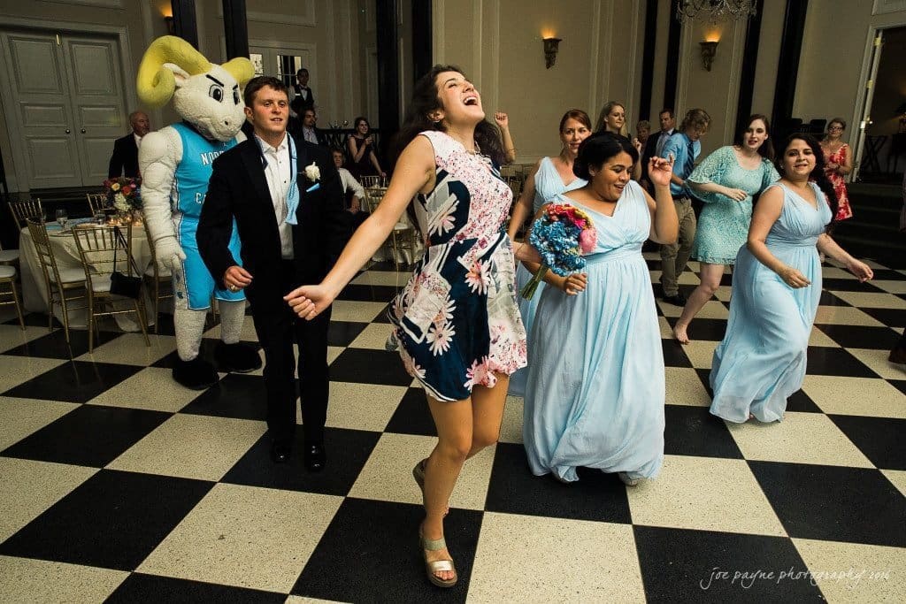 duke chapel & carolina inn wedding - sidney & andrew A group of people, including a woman in a floral dress and bridesmaids in blue dresses, are dancing on a black and white checkered floor. A mascot is in the background, and the scene takes place in a well-lit room with elegant decor.