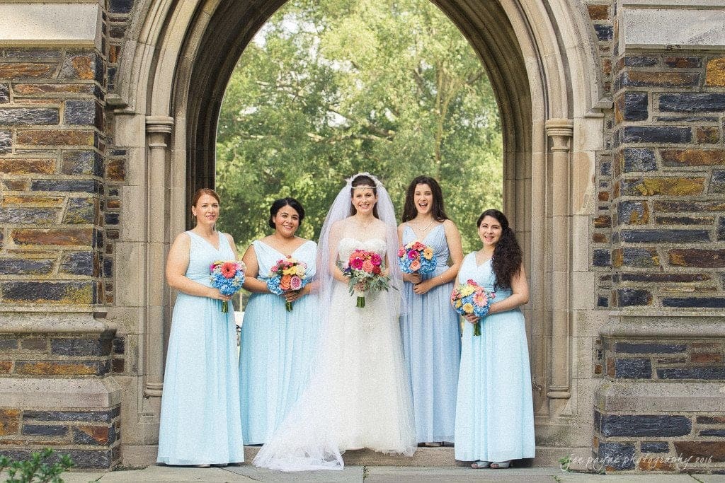 duke chapel & carolina inn wedding - sidney & andrew A bride in a white dress and veil stands with four bridesmaids in light blue dresses. They are holding colorful bouquets and are positioned under a stone arch with green trees in the background.