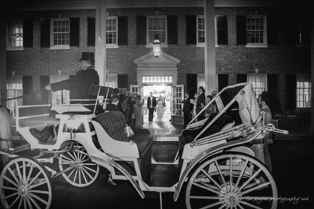 duke chapel & carolina inn wedding - sidney & andrew A black and white image of a horse-drawn carriage parked outside a large brick building, possibly at night. People stand on the porch, illuminated by lights from within the building. The scene suggests a formal event or celebration.