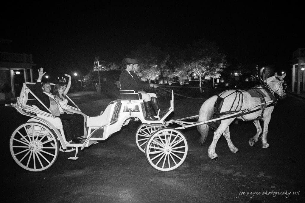 duke chapel & carolina inn wedding - sidney & andrew A black and white photo of a horse-drawn carriage at night. Two people wave from the carriage as a driver steers the horse along a road. Trees and buildings are softly illuminated in the background.