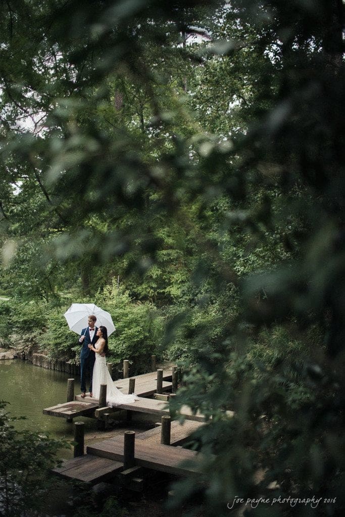 A couple stands on a wooden dock by a tranquil pond, surrounded by lush greenery. They share an umbrella, adding a romantic touch to the serene, natural setting.
