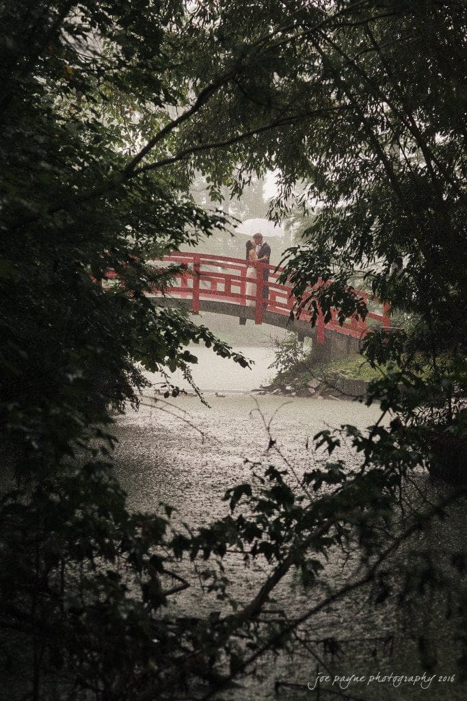 A couple embraces on a red arched bridge amidst lush greenery. Its a rainy day, with water rippling below and leaves gently framing the scene.
