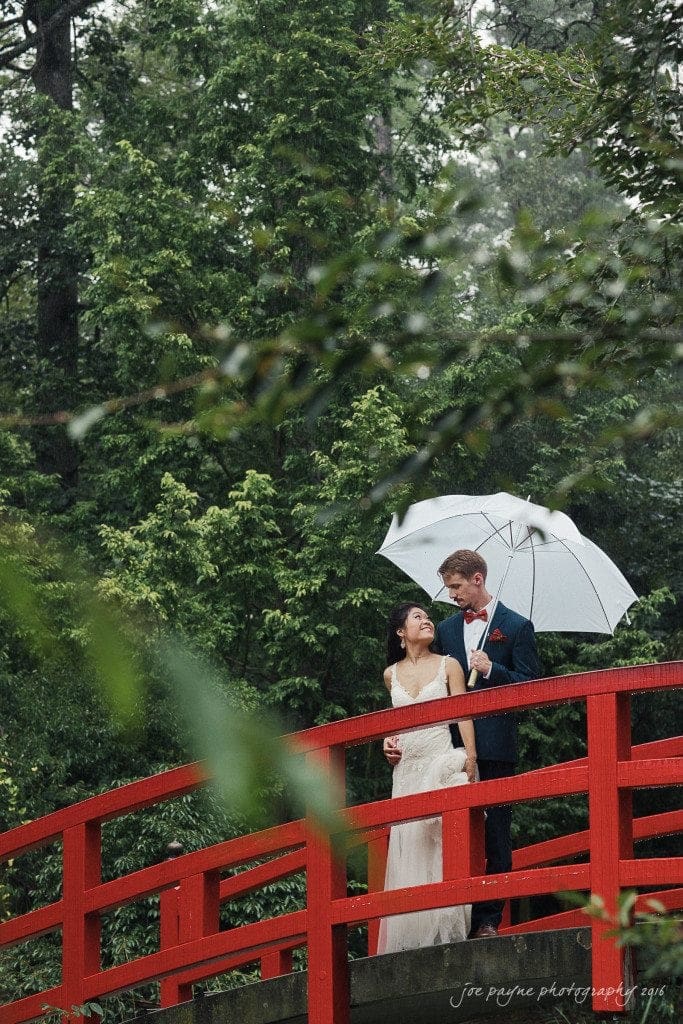 A couple stands on a red bridge in a lush green forest. The woman in a white dress smiles while the man in a dark suit holds a white umbrella.