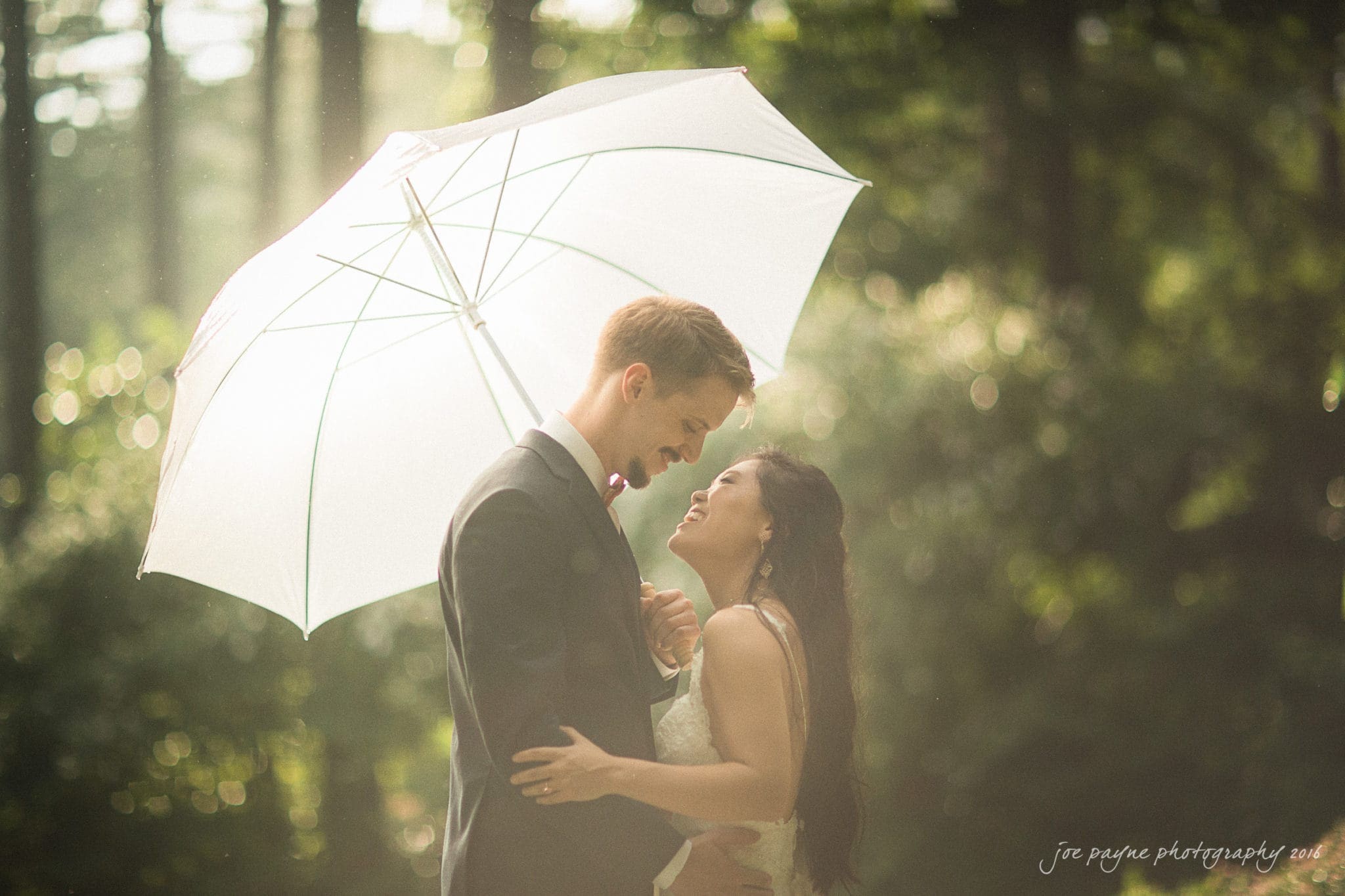 A couple in formal attire stands under a white umbrella in a sunlit forest. They smile at each other affectionately, creating a romantic and serene atmosphere.