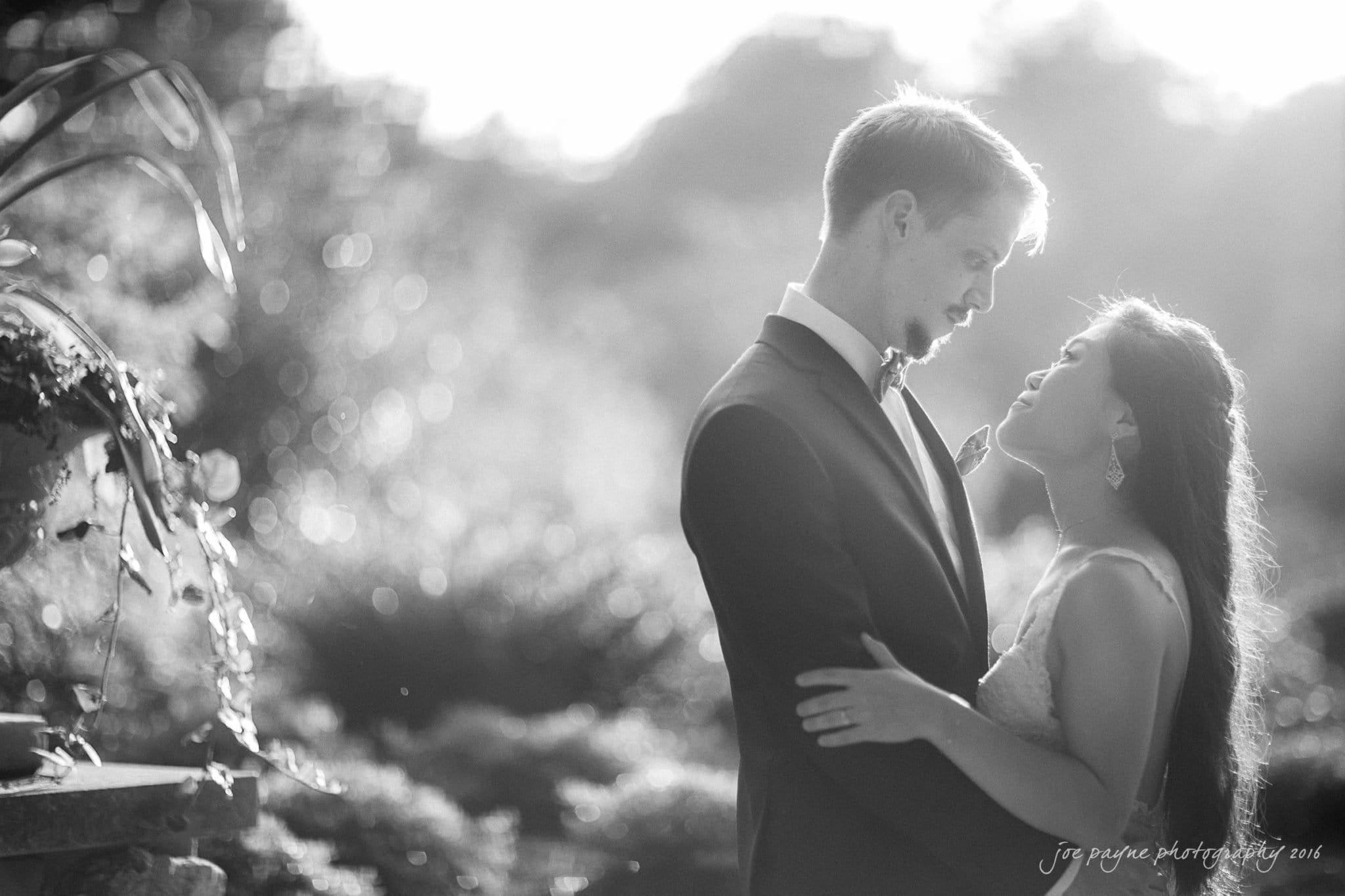 A black and white photo of a couple embracing outdoors, with the groom in a suit and the bride in a wedding dress. They look at each other lovingly, surrounded by soft, blurred foliage in the background.