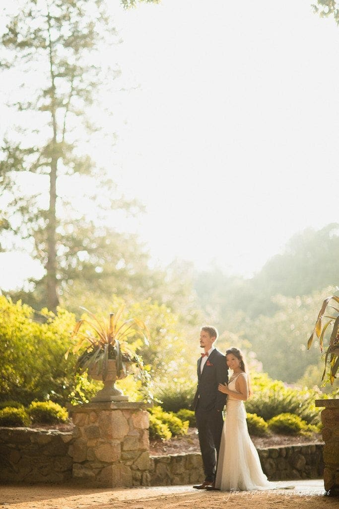 A bride and groom stand outdoors in a sunlit garden. The groom is in a dark suit with a red bow tie, and the bride is wearing a white gown. They are surrounded by lush greenery and stone planters, with sunlight streaming through the trees.
