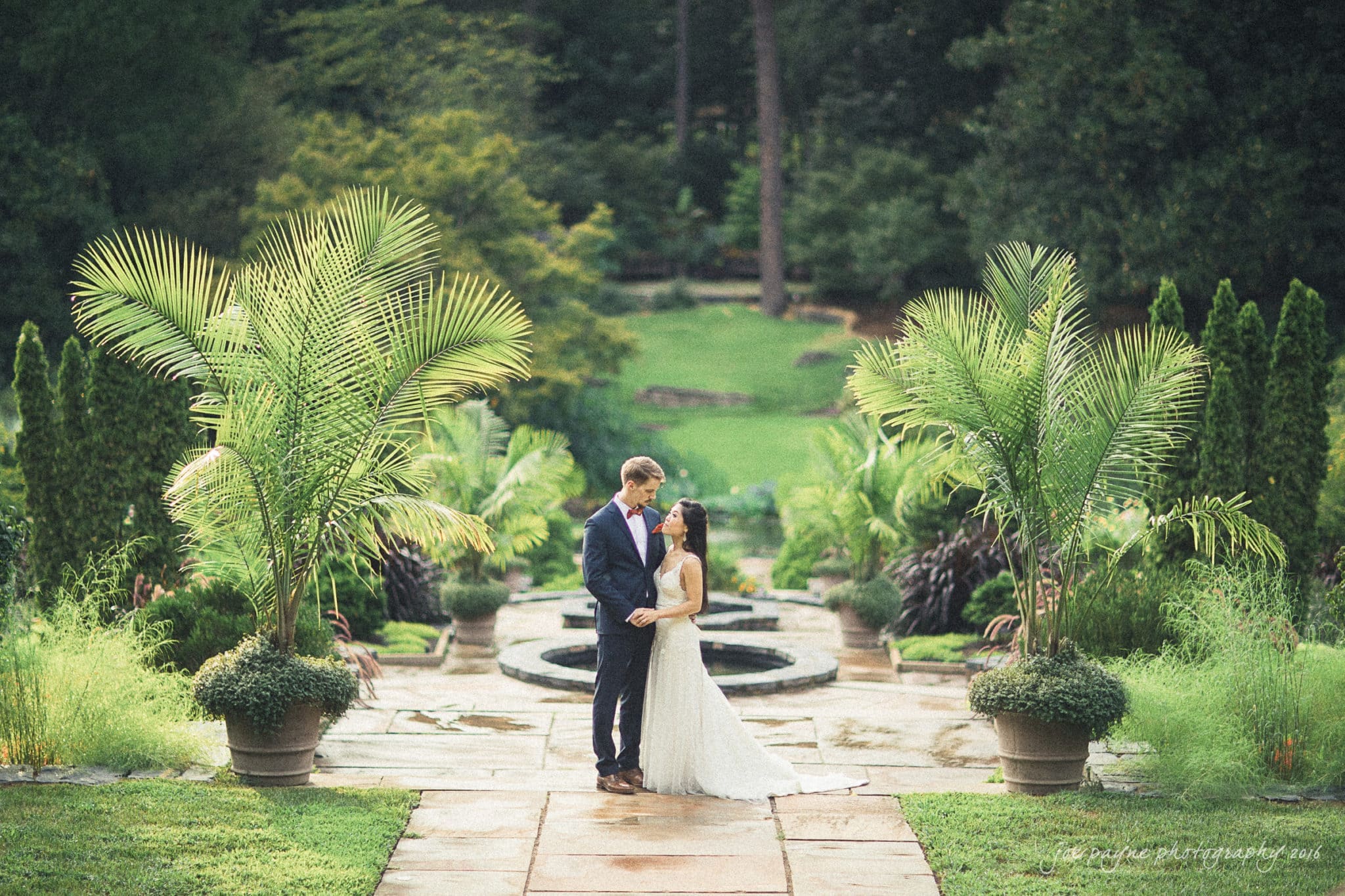 A couple stands lovingly in an elegant garden setting, surrounded by lush greenery and tall palm plants. A circular fountain in the background adds to the romantic atmosphere. The pathway is lined with neatly trimmed shrubs.