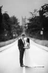 A couple in wedding attire stands on a deserted road, surrounded by trees. They gaze at each other lovingly as the blurred background showcases a tall building. The image is in black and white, creating an elegant, timeless feel.