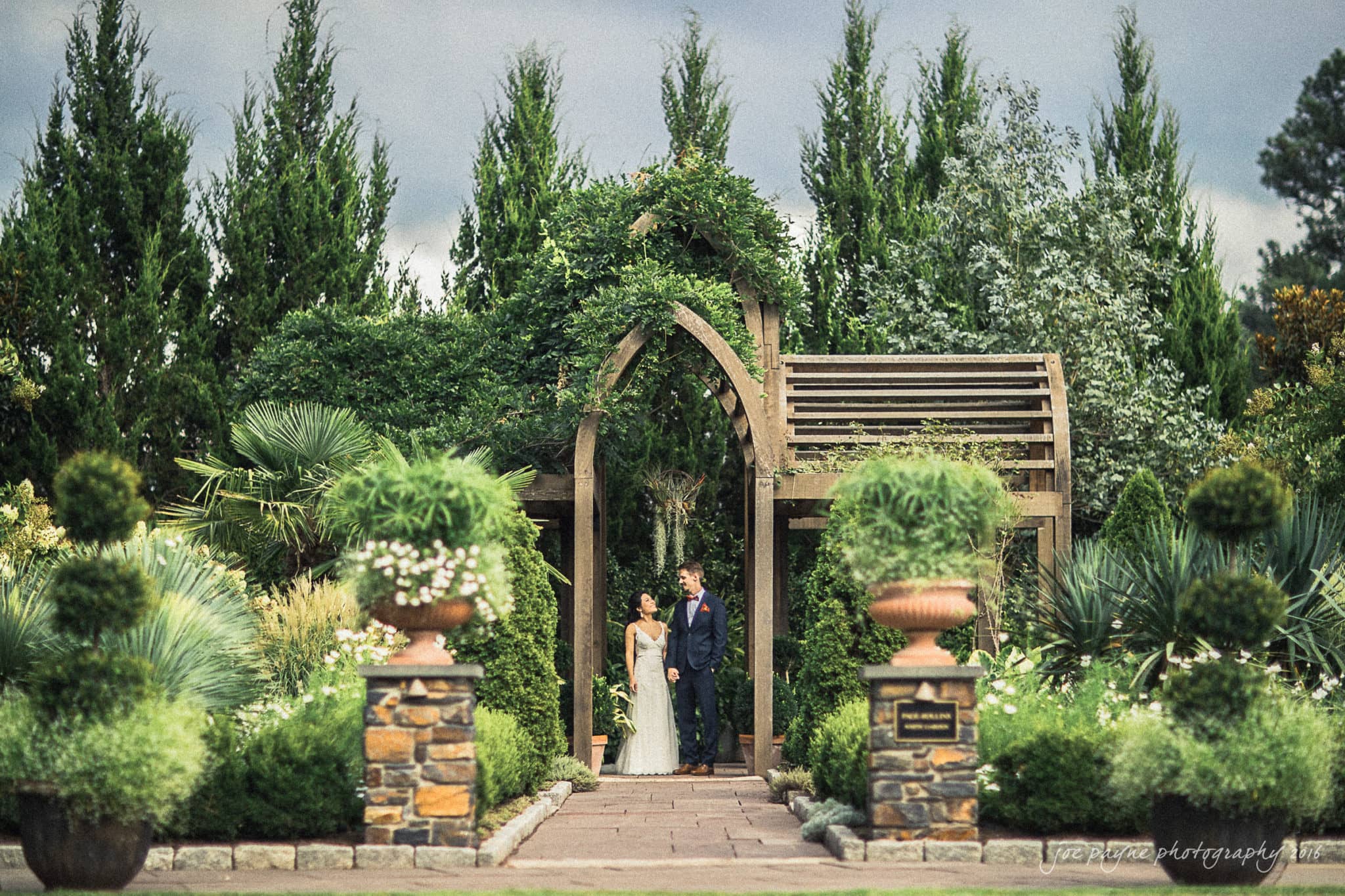 A couple stands in a lush garden under a wooden archway surrounded by green trees and plants. They are dressed in formal attire, with the background featuring tall foliage and cloudy skies, creating a serene and romantic atmosphere.