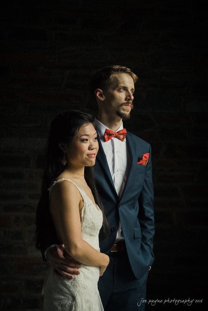 A bride and groom stand closely together in formal attire, with the groom wearing a suit and bow tie and the bride in a white lace dress. They are posed against a dark backdrop, gazing into the distance.