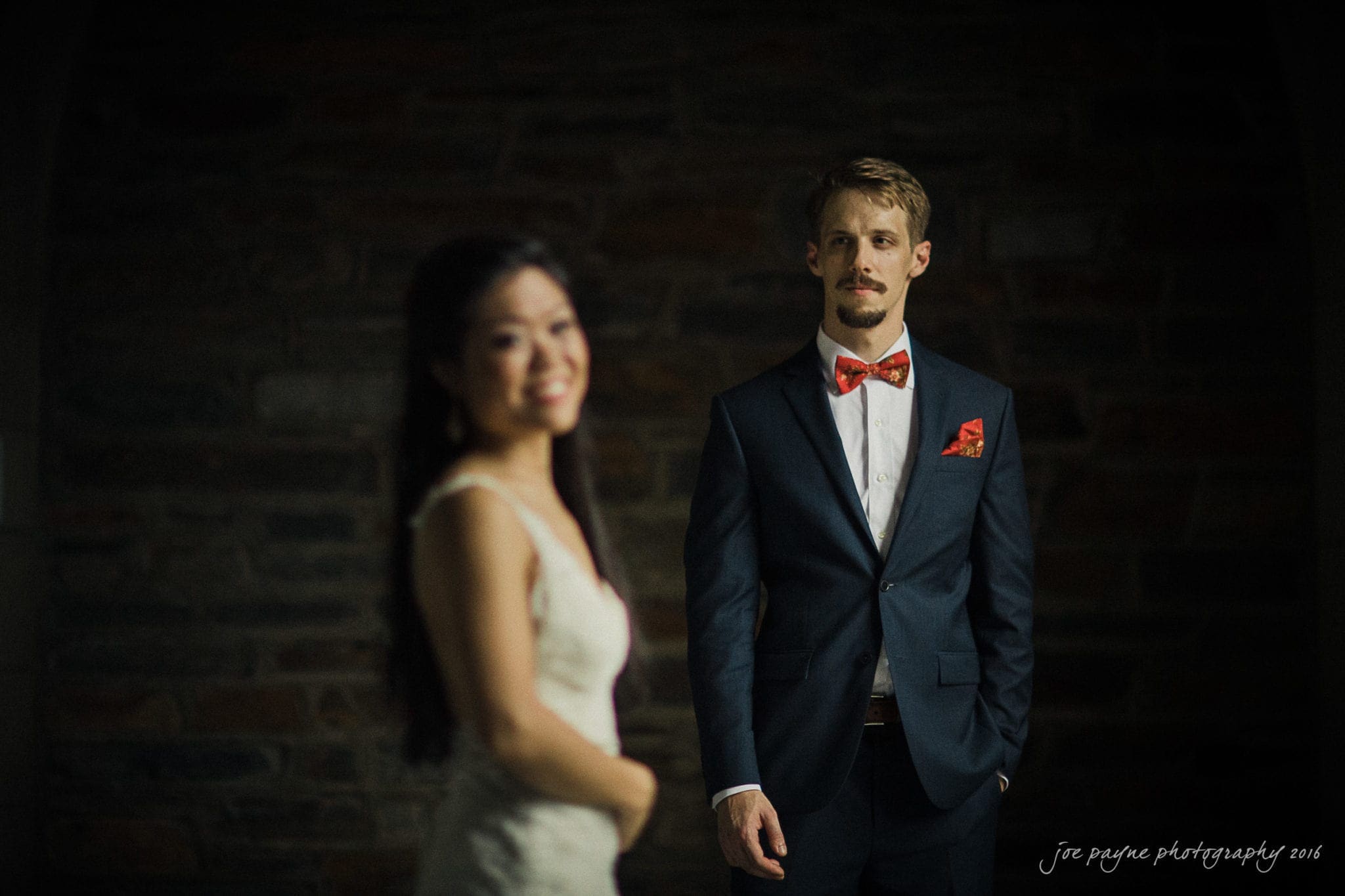 A man in a dark suit and red bow tie stands against a dark brick background, looking forward. A woman in a white dress stands in the foreground, slightly out of focus, looking towards him with a smile.